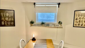 A dining area with a wooden table, white chairs, a window with a view, and a shelf with plants and jars. - Cozy Studio - Long Term 1 Month + Only (Saginaw)