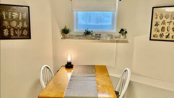 Un coin repas avec une table en bois, des chaises blanches, une fenêtre donnant sur un beau paysage et une étagère ornée de plantes et de bocaux.