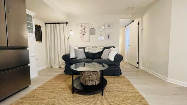 A modern living room with a dark sofa, a round coffee table, and a kitchen area with a refrigerator. - Cozy Studio - Long Term 1 Month + Only (Saginaw)
