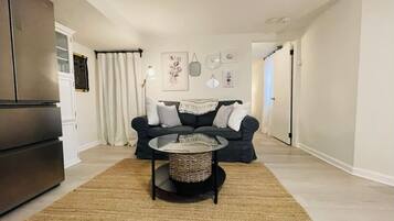 Un salon moderne avec un canapé foncé, une table basse ronde et un coin cuisine équipé d’un réfrigérateur.