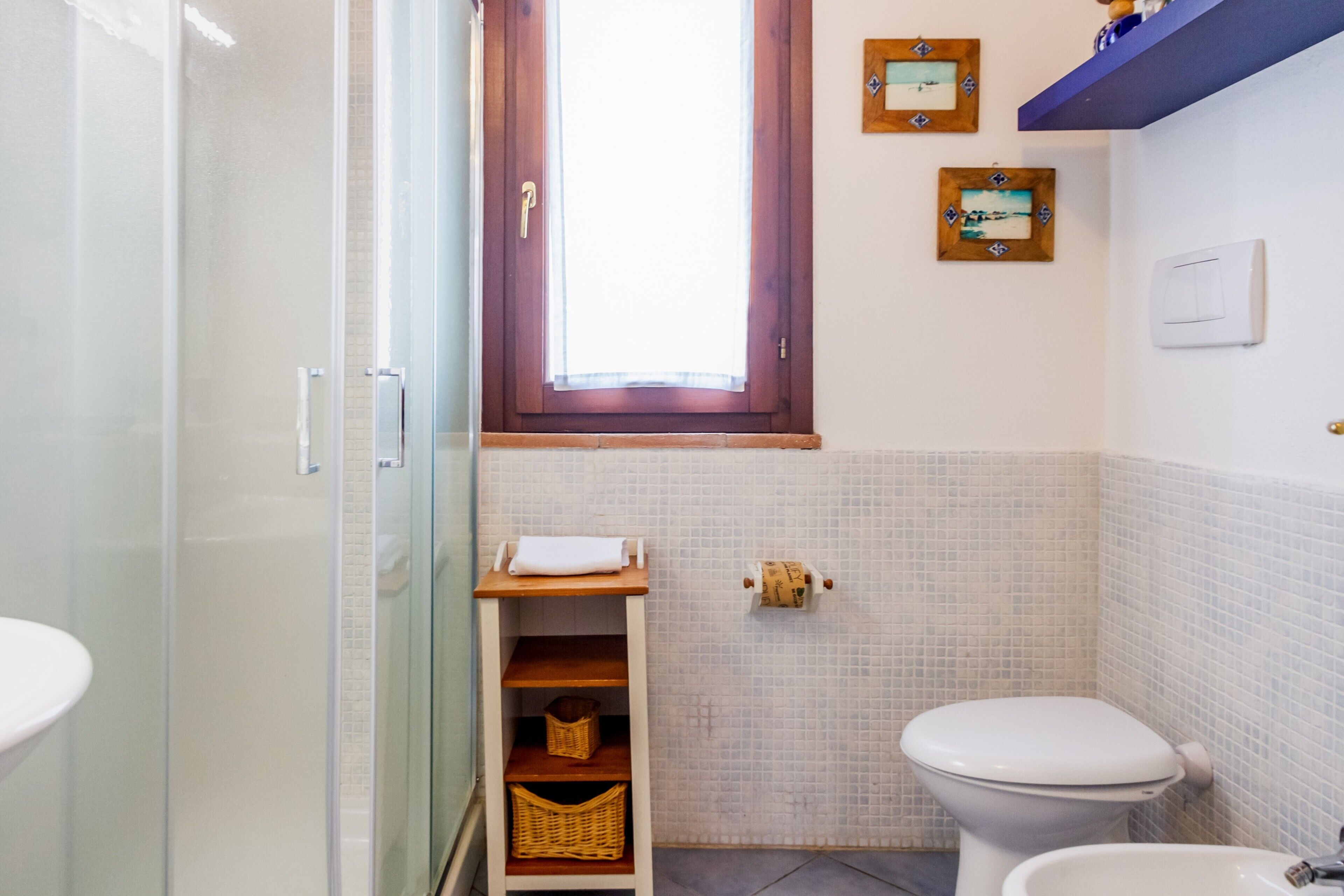 A bathroom with a glass shower enclosure, a toilet, a bidet, a small shelf with baskets, and a window with a wooden frame.