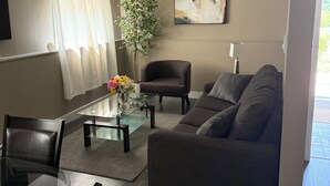 A modern living room with a glass coffee table, a grey sofa, a dark armchair, and a dining area with a glass table and chairs. - Cozy 2-bedroom retreat – comfort, style, and relaxation await you (Bedford)