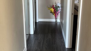 A hallway with dark wood flooring, beige walls, and a ceiling light fixture. There is a painting on the left wall and a vase with flowers on the right wall. - Cozy 2-bedroom retreat – comfort, style, and relaxation await you (Bedford)