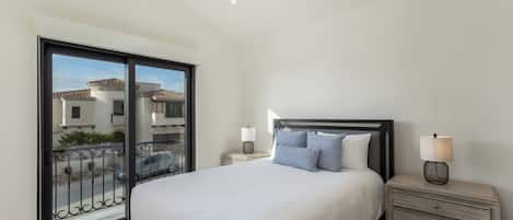 A bedroom with a large bed, a nightstand, a lamp, and a view of the outside through a sliding door.