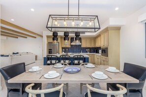 A modern dining area with a wooden table set for six, surrounded by black chairs. A large rectangular chandelier hangs above, and the room features a kitchen with light-colored cabinets and a blue herringbone backsplash.