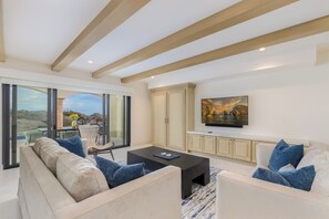 A modern living room with a large sofa, a coffee table, and a TV mounted on the wall. There is a sliding glass door leading to an outdoor area with a view of the landscape.
