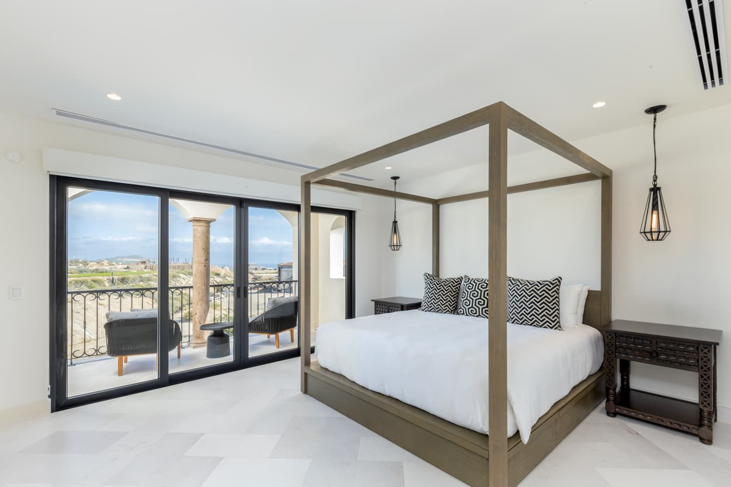 A bedroom with a four-poster bed, a large sliding glass door leading to a balcony, and a view of the landscape.
