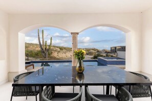 A dining area with a black table and chairs, a vase with flowers, and a view of a desert landscape with cacti and a pool.
