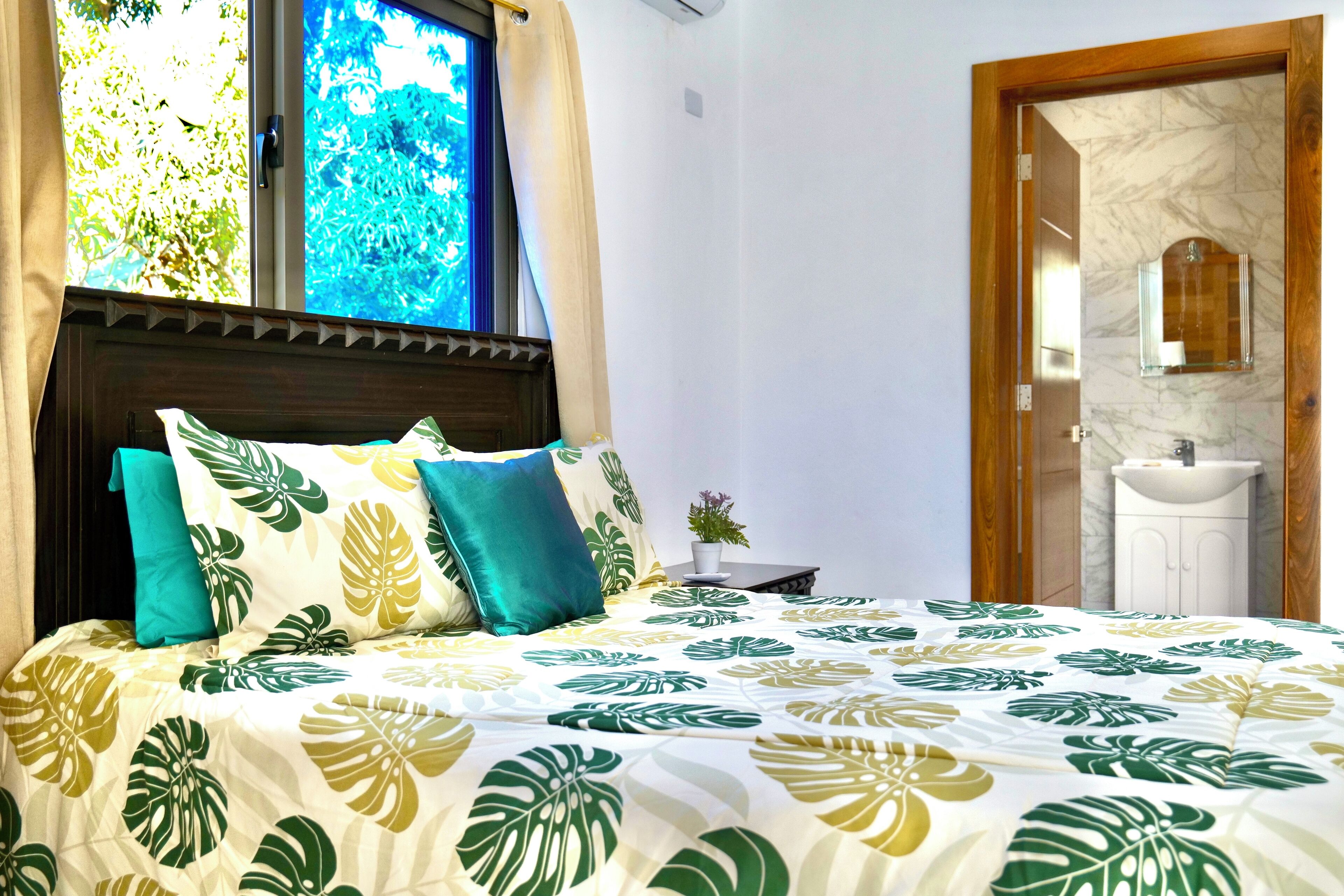 A bedroom with a bed, a window with curtains, a bedside table with a plant, and a bathroom with a sink and mirror.