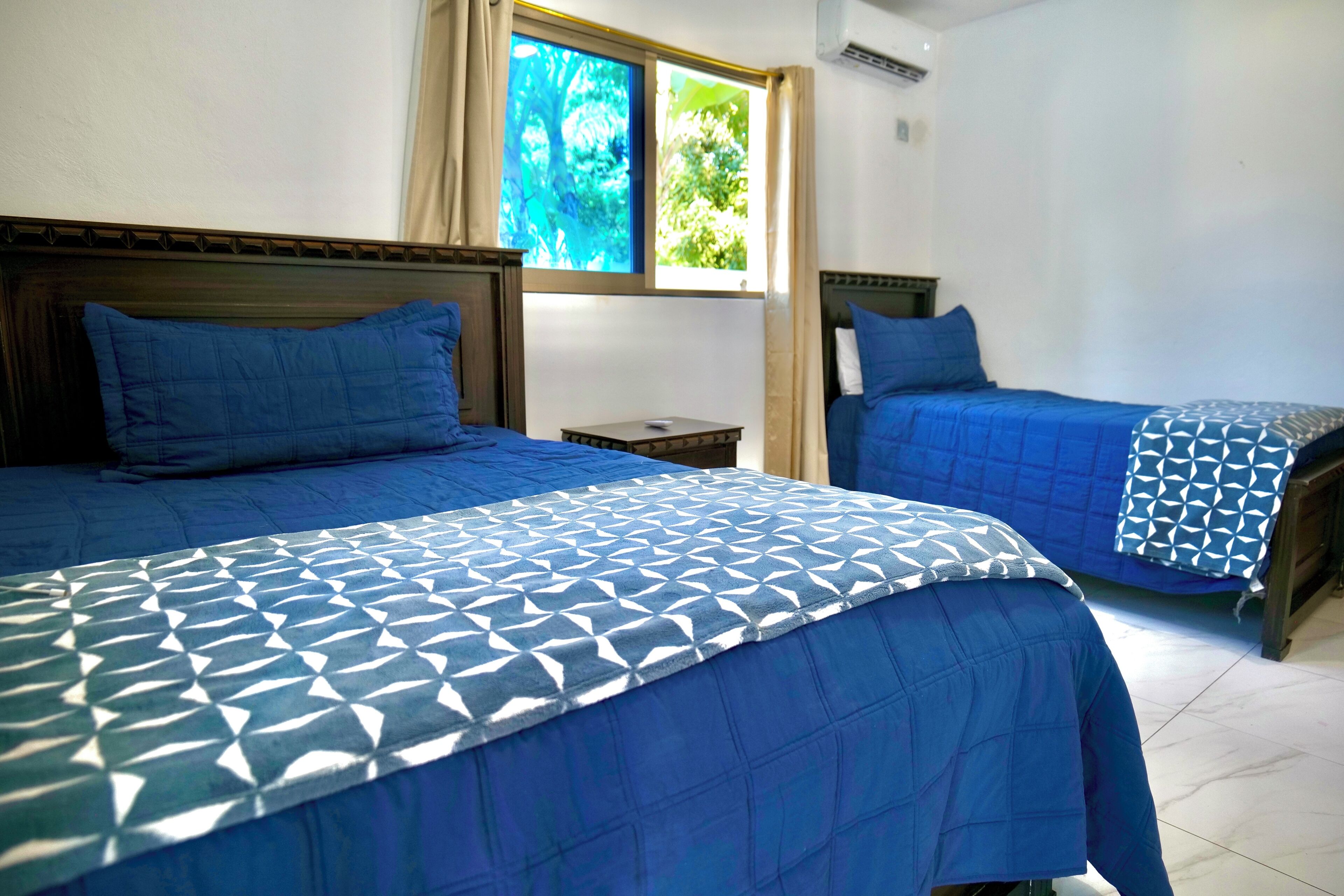 A room with two beds, each with a blue bedspread and matching pillows. There is a window with a view of greenery outside.