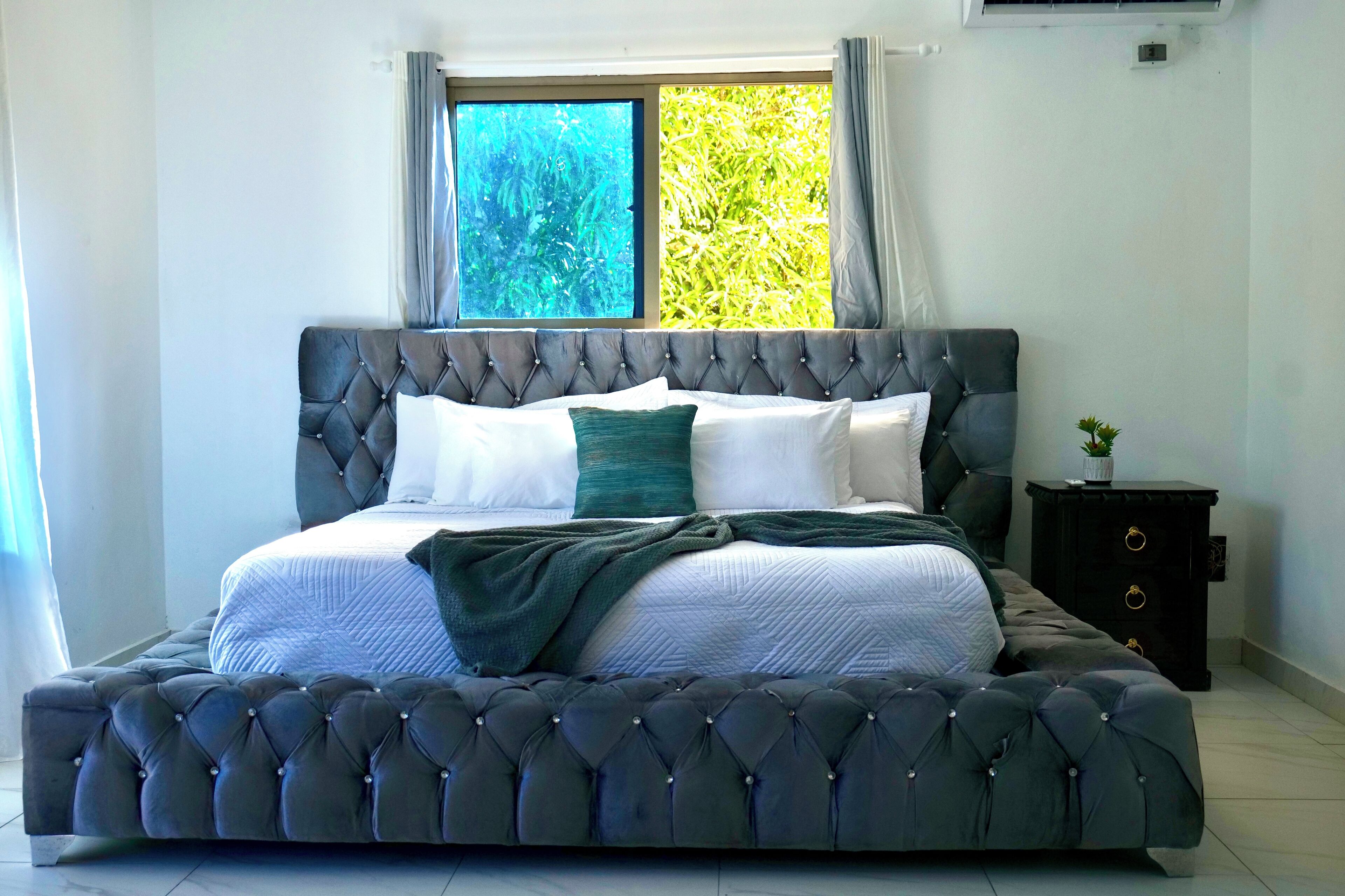 A large bed with a tufted headboard, a dark nightstand with drawers, and a window with curtains.