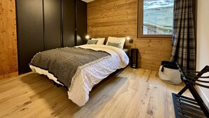 A bedroom with a wooden floor, a bed with a white and gray duvet, a nightstand, a window with a view of a snowy landscape, and a plaid curtain. - Cozy T2 and mountain area, a stone's throw from the slopes! (CHATEL)