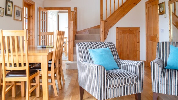 A spacious living area with a wooden dining table and chairs, a striped armchair with a blue pillow, and a wooden staircase. - Creagan by Interhome (North Skye)
