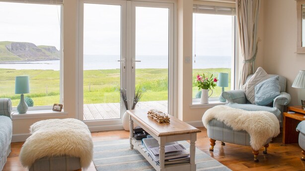 Ein Zimmer mit großem Fenster und Blick auf Meer und Klippen, mit Sofa, Sessel, kleinem Tisch mit Lampe und einer dekorativen Herz-Form an der Wand.