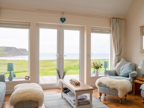 Habitación con un amplio ventanal que ofrece vistas al mar y a los acantilados, un sofá, una silla, una mesita con una lámpara y un corazón decorativo en la pared.