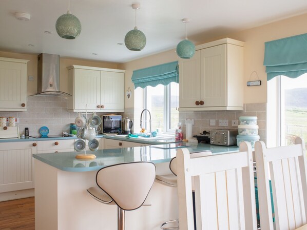 Una cocina moderna con armarios blancos, una isla central con taburetes altos y lámparas colgantes en forma de globos verdes, y vistas al exterior.