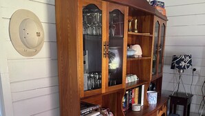 A wooden cabinet with glass doors, containing various items like books, a lamp, and kitchenware. - Quintessential Tassie Shack (South Mount Cameron)