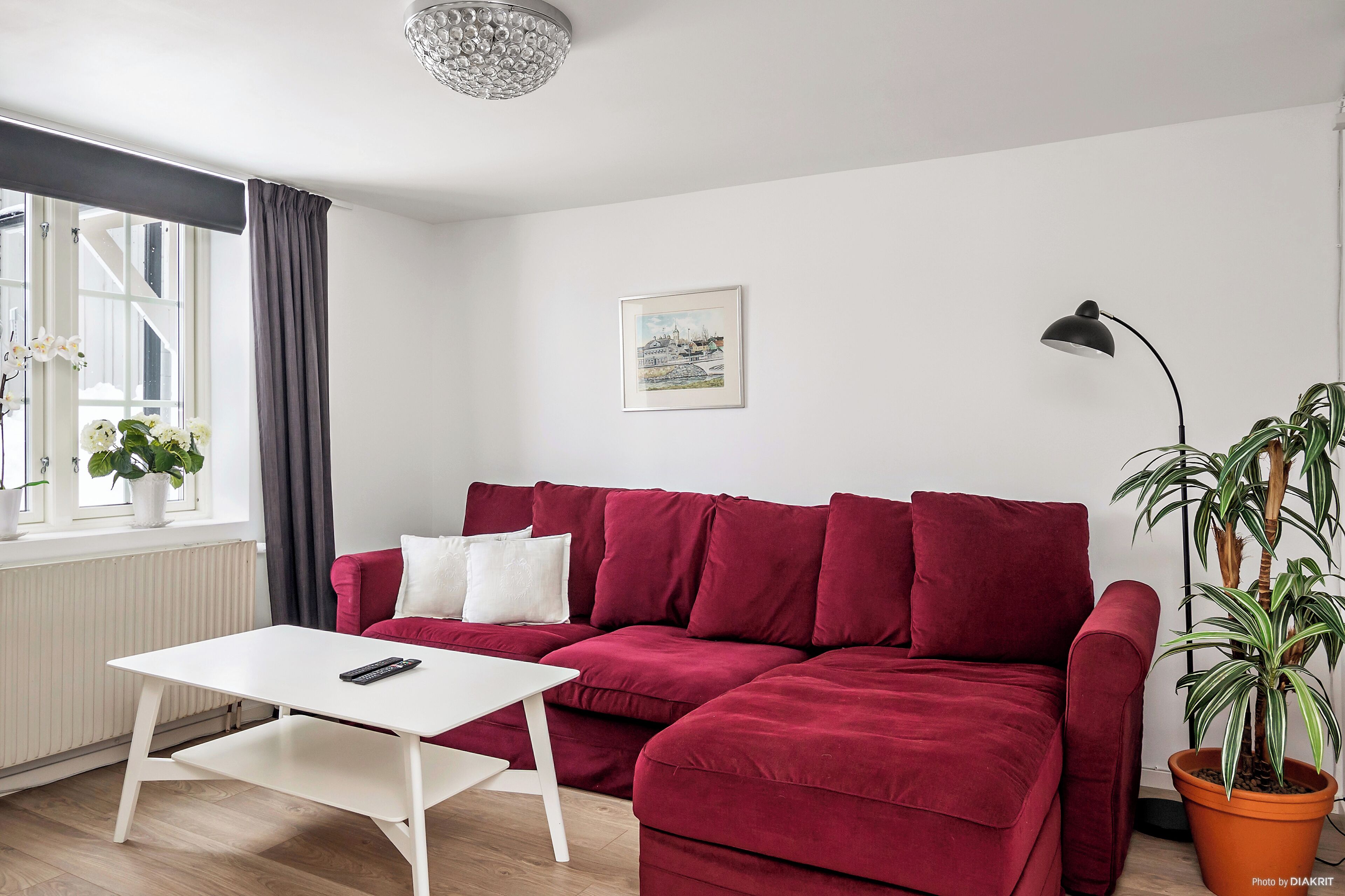 En rød sofa, et hvidt bord og en potteplante i et rum med et vindue, en radiator og et billede i ramme på væggen.