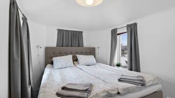 A neatly made bed with a patterned bedspread and pillows, grey curtains, and a bedside plant.