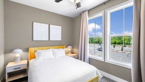 Une chambre d’hôtel comprenant un lit, deux tables de chevet, un ventilateur de plafond et de grandes fenêtres donnant sur un parking et des espaces verts.