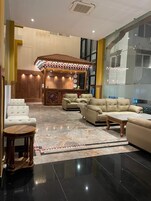 A spacious lobby with a central seating area featuring sofas and a coffee table, a decorative wall with clocks, and a large window providing a view of the outside.
