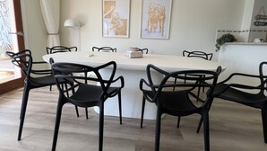 A dining area with a white table and black chairs, two framed artworks on the wall, and a kitchen area with a plant.