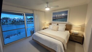 A hotel room with a large bed, a nightstand, a ceiling fan, a window with a view of a body of water and buildings, and a framed picture on the wall.