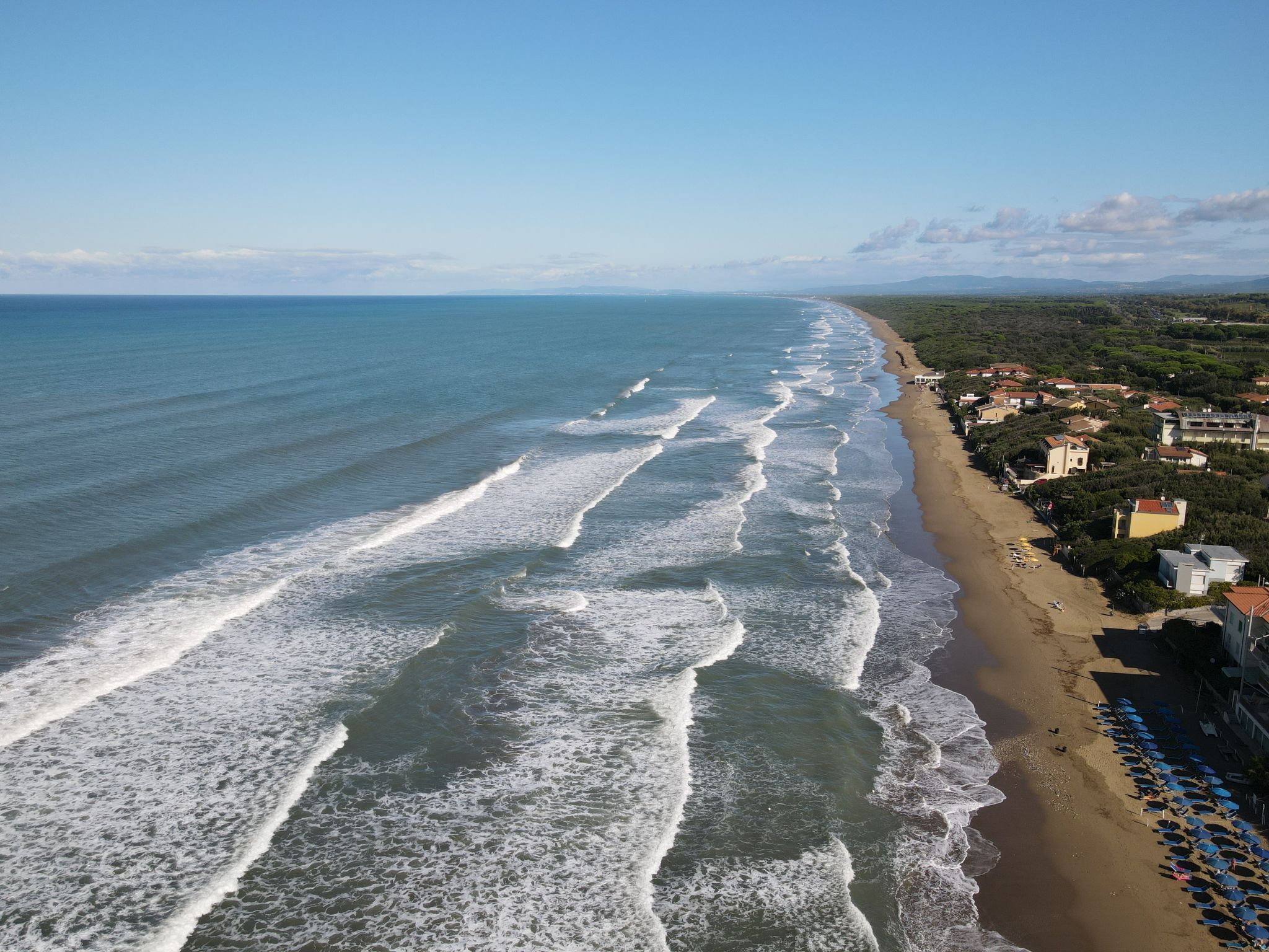An aerial view of a beach with waves, a row of houses, and a clear sky.