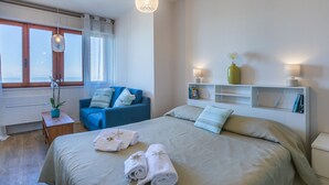Un dormitorio moderno con una cama grande, un sillón azul, una mesita de noche con lámpara y vista al mar a través de la ventana.