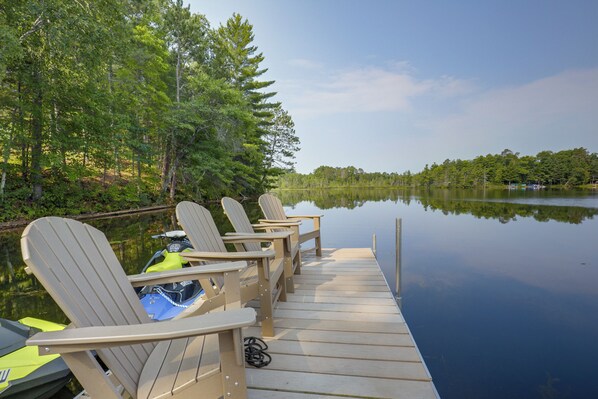 Eine Holzsteg mit mehreren Adirondack-Stühlen und einem Jetski, mit Blick auf einen ruhigen, von Bäumen gesäumten See.