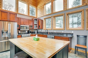A modern kitchen with wooden cabinets, stainless steel appliances, a large island, and large windows offering a view of snow-covered trees.