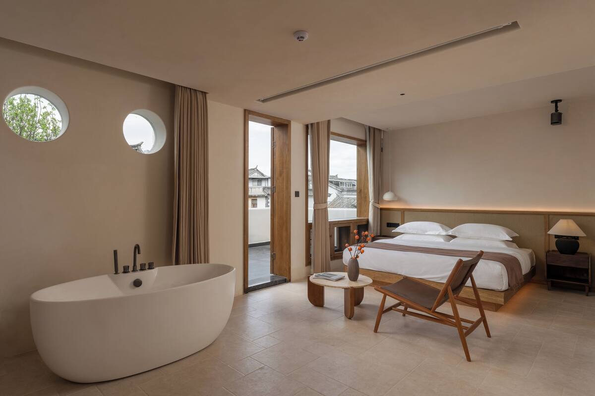 Un hotel moderno con una bañera grande, una cama con ropa blanca, una mesita redonda con un florero y vistas a un edificio tradicional por la ventana.