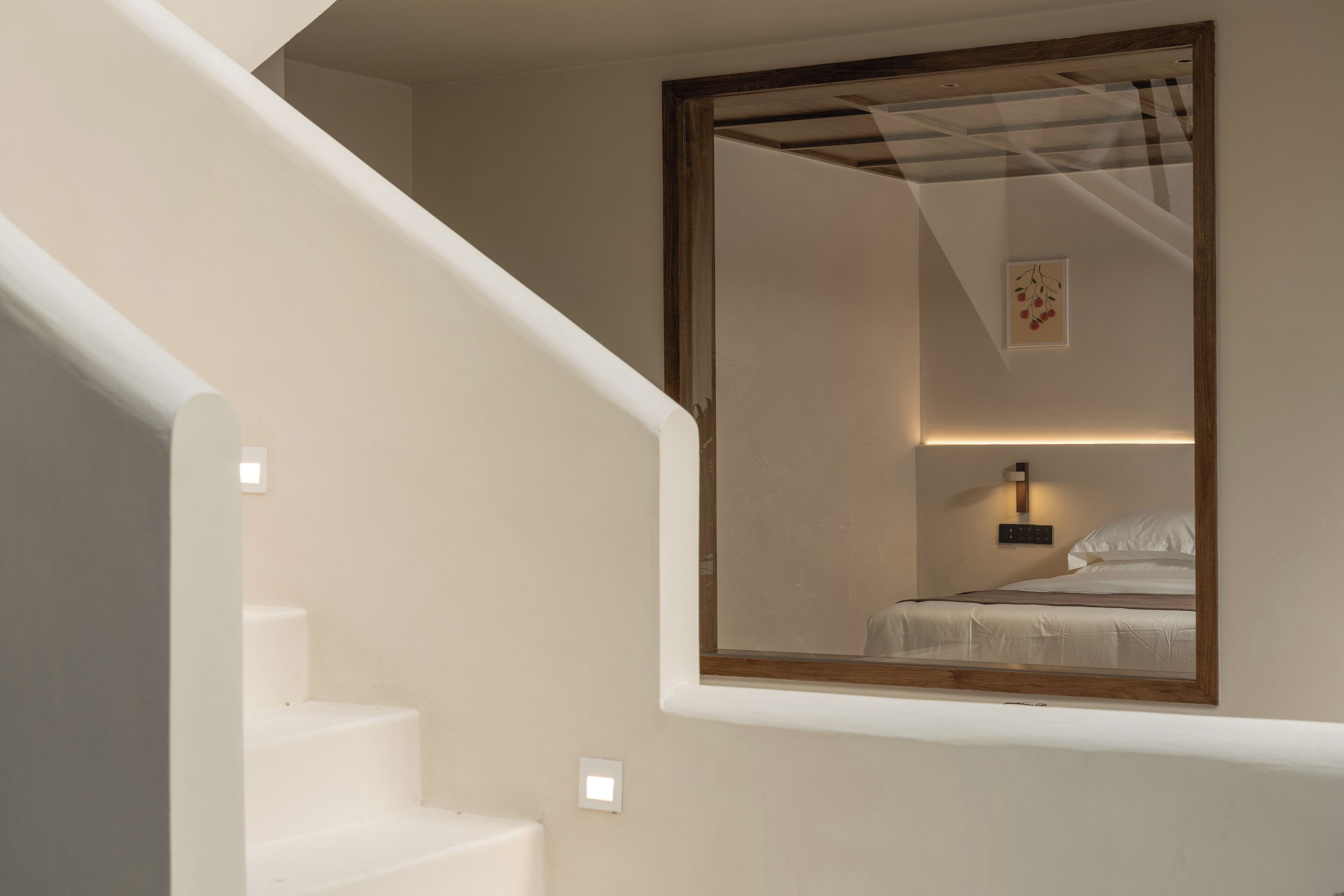 Una escalera con barandilla blanca conduce a un dormitorio con un espejo grande que refleja una cama y un aplique de pared.