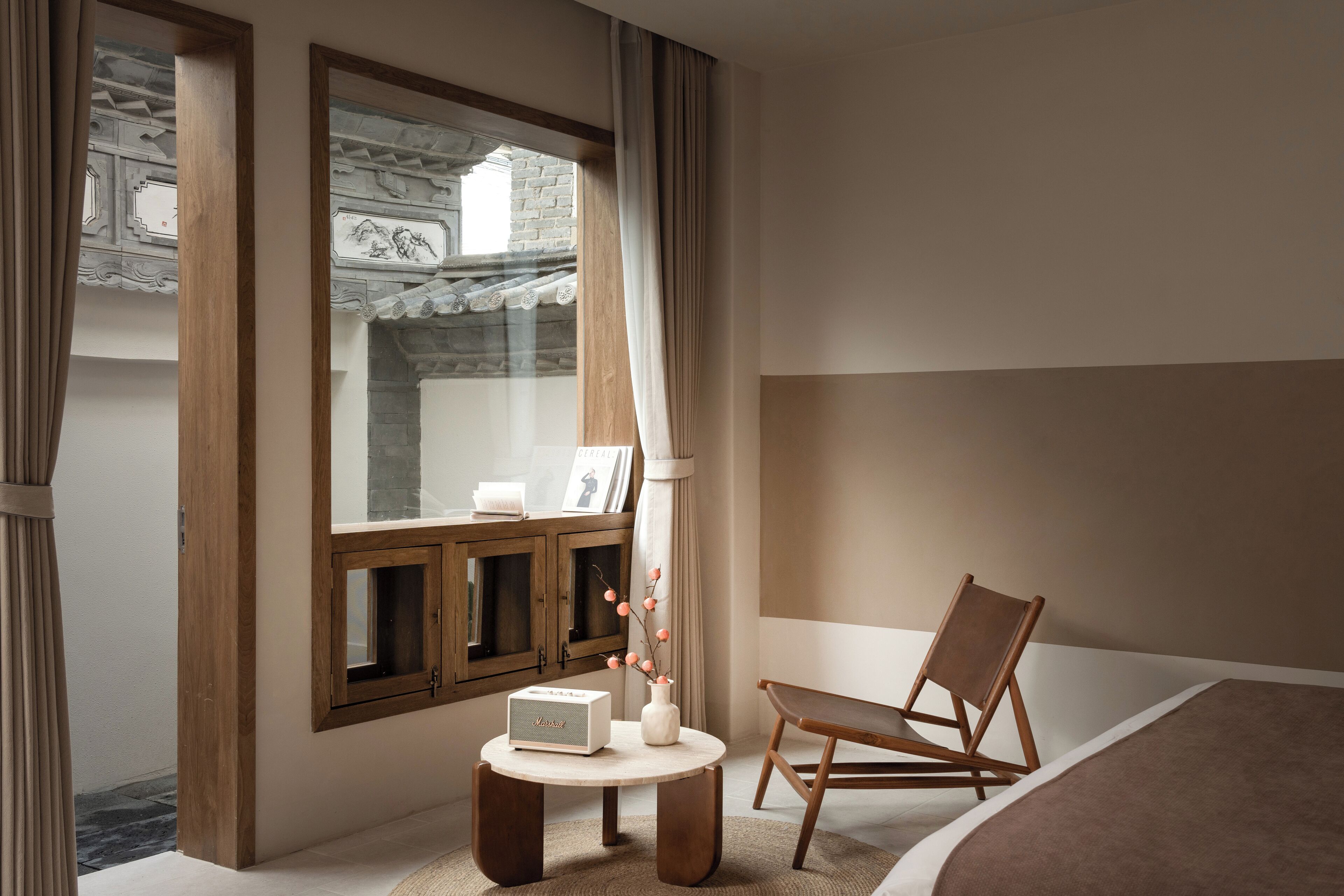 A hotel room with a large window, a wooden chair, a small round table with a radio, and a bed with a brown blanket.