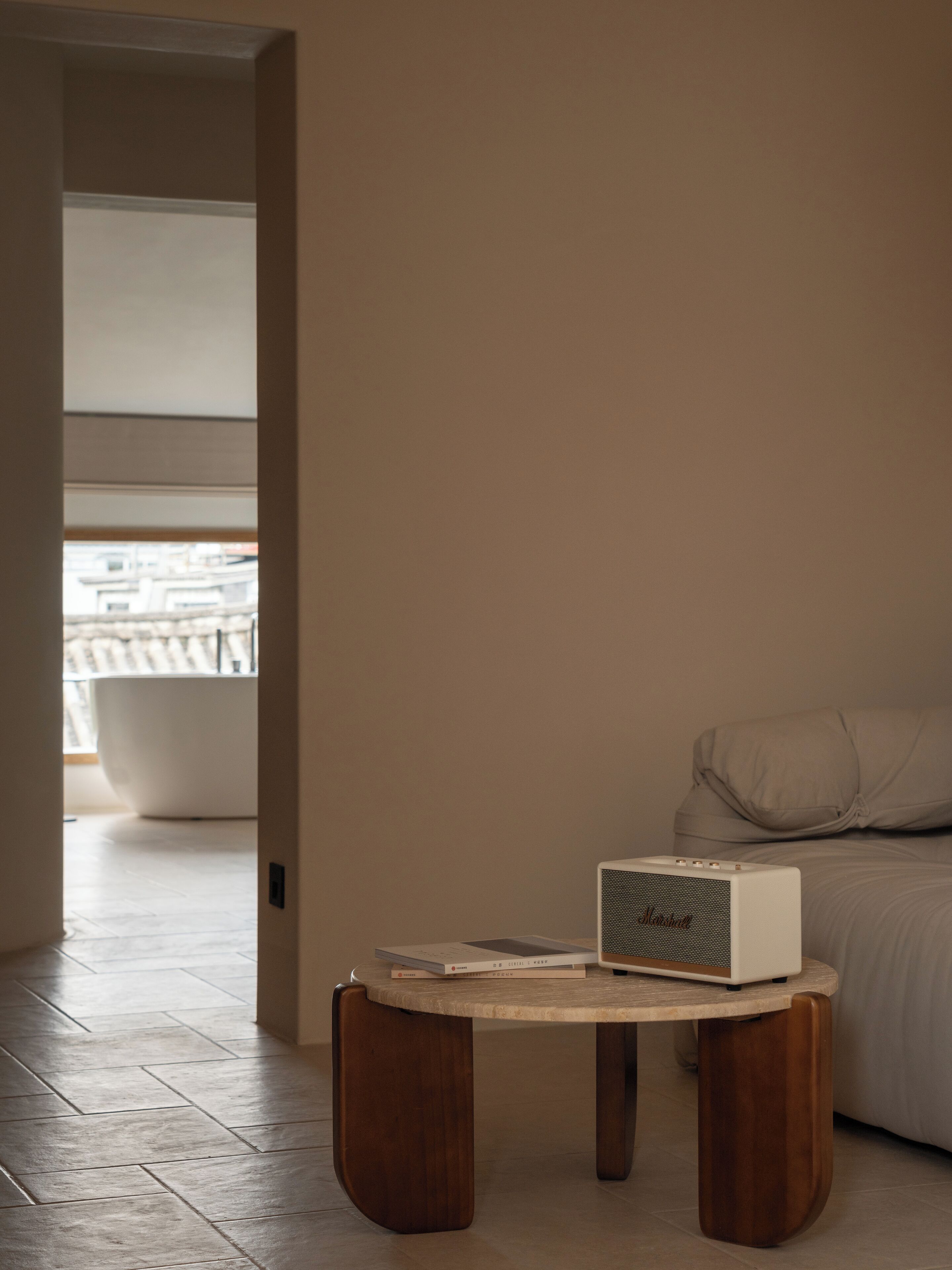 Une pièce moderne au design minimaliste, avec une table ronde en bois sur laquelle sont posés un haut-parleur blanc et des livres, une grande fenêtre offrant une vue sur les bâtiments et, au fond, une grande baignoire blanche.