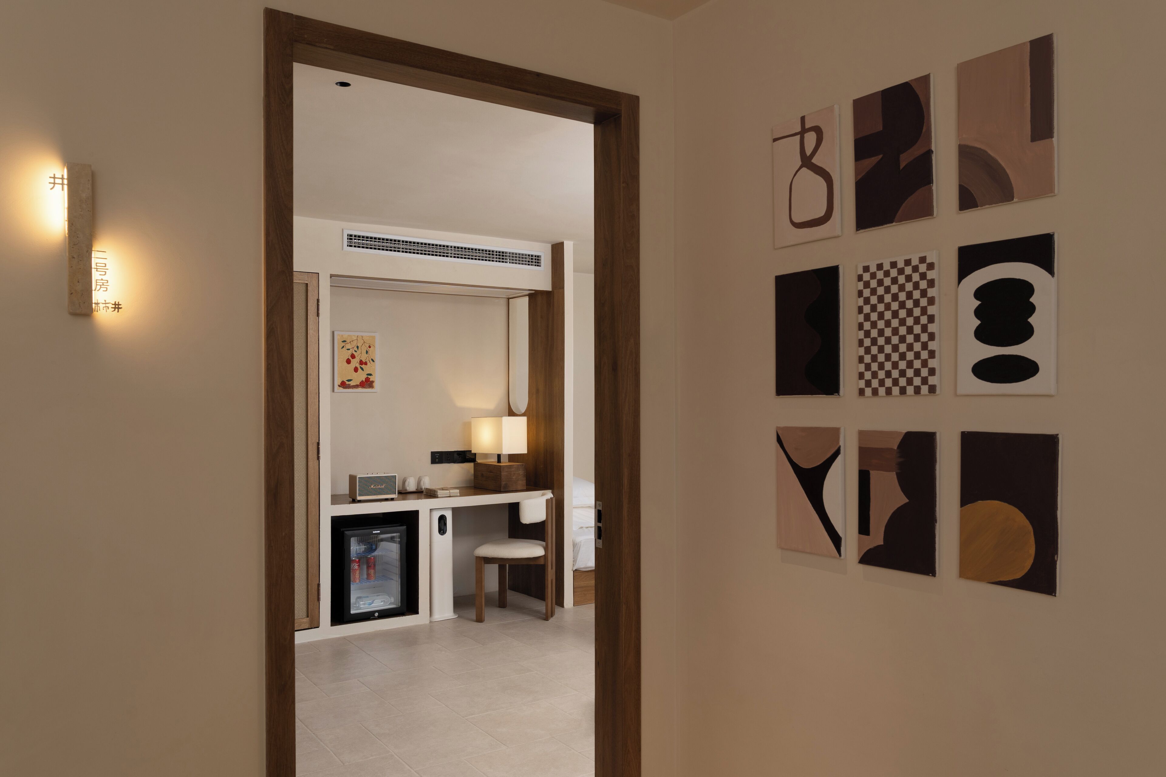 A hallway leading to a room with a desk, chair, and a refrigerator. There are wall-mounted artworks and a wall-mounted light fixture.