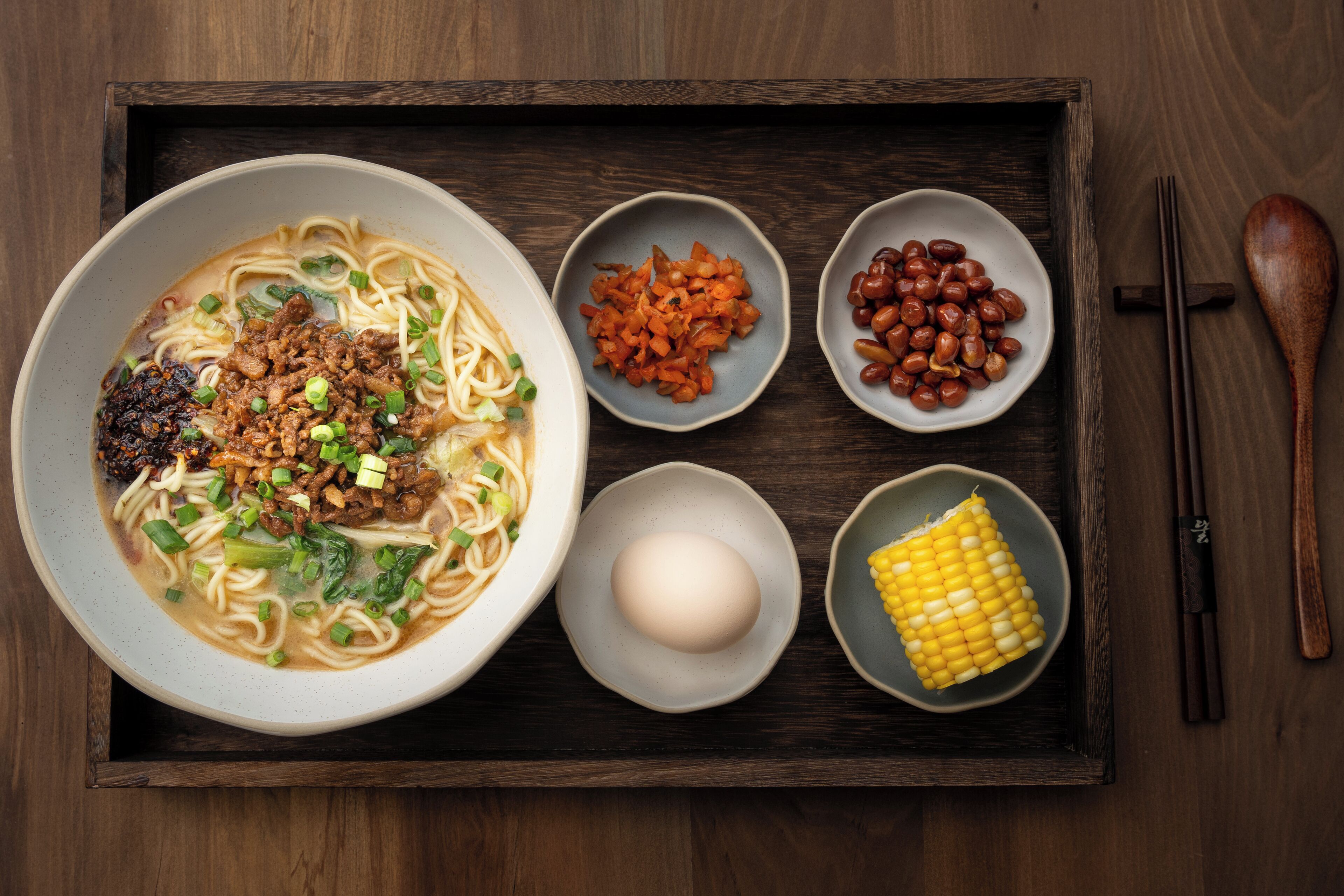 A tray with a bowl of ramen noodles topped with meat and green onions, accompanied by small bowls of pickled vegetables, peanuts, a boiled egg, and a corn cob.