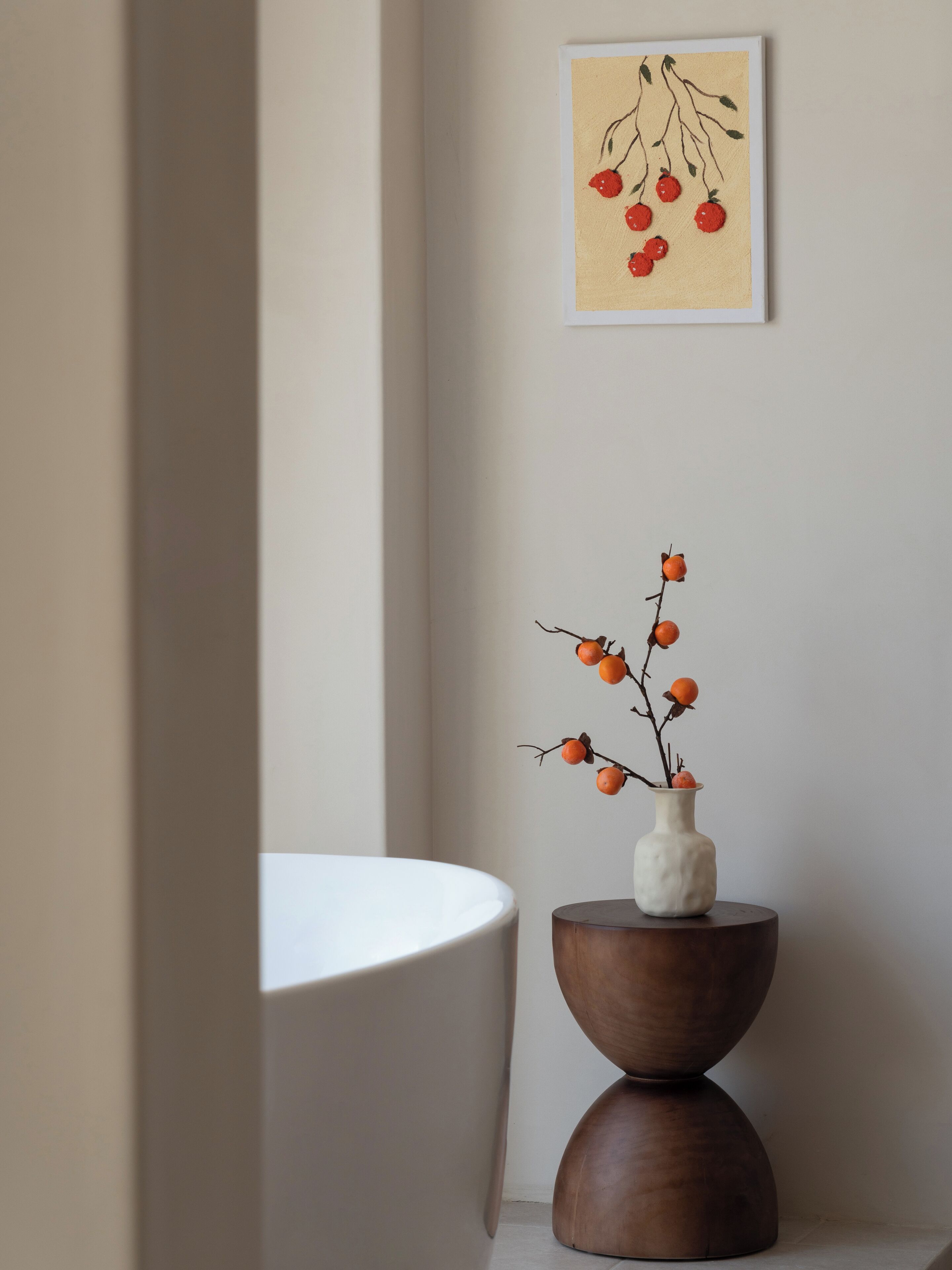 A bathroom with a white bathtub, a wooden side table with a vase of orange berries, and a framed picture on the wall.
