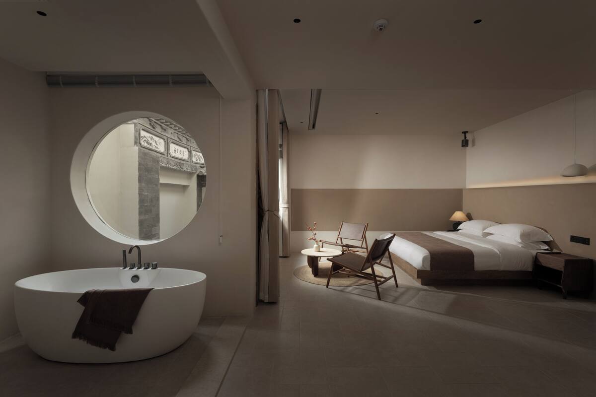 Un dormitorio moderno con un espejo redondo grande, una bañera, una cama y una silla.
