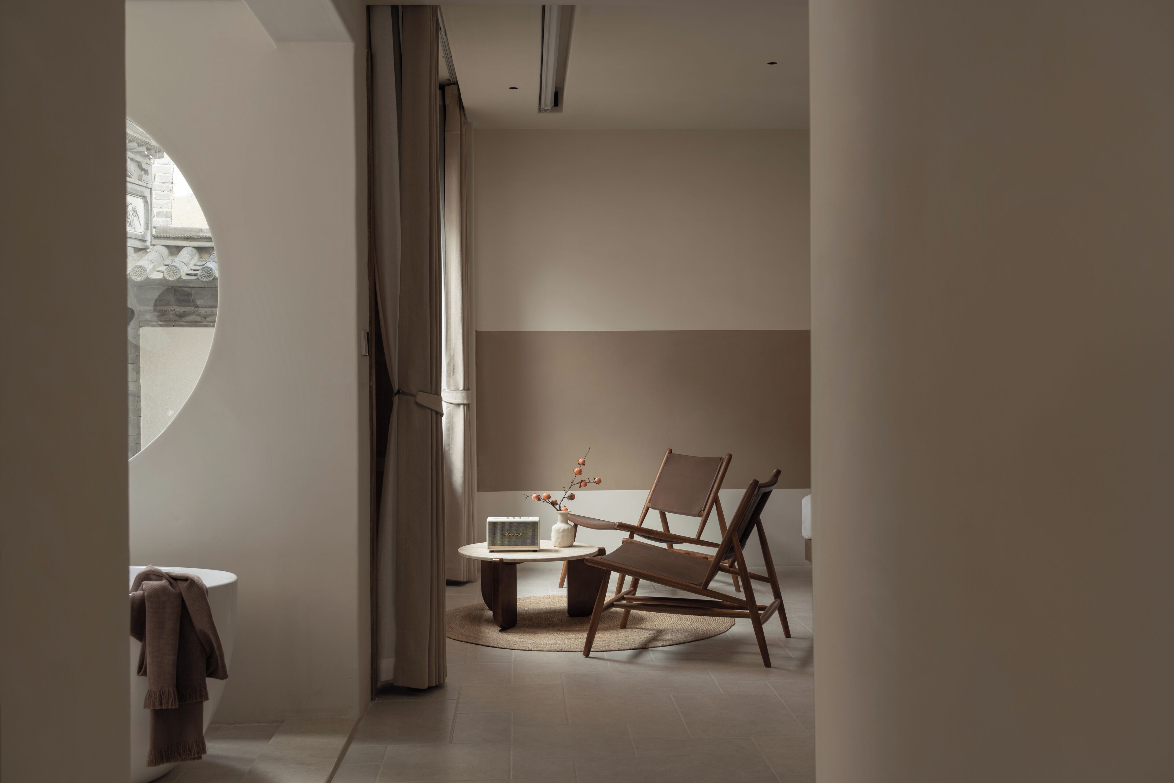 A modern room with a round window, a wooden chair, a small table, and a white vase with flowers.