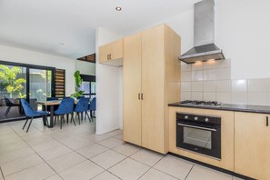 A modern kitchen with wooden cabinets, a gas stove, and an oven. There is a dining area with blue chairs and a table, and a large window providing natural light. - Dolphin Dreamin' with magnesium pool (Agnes Water)
