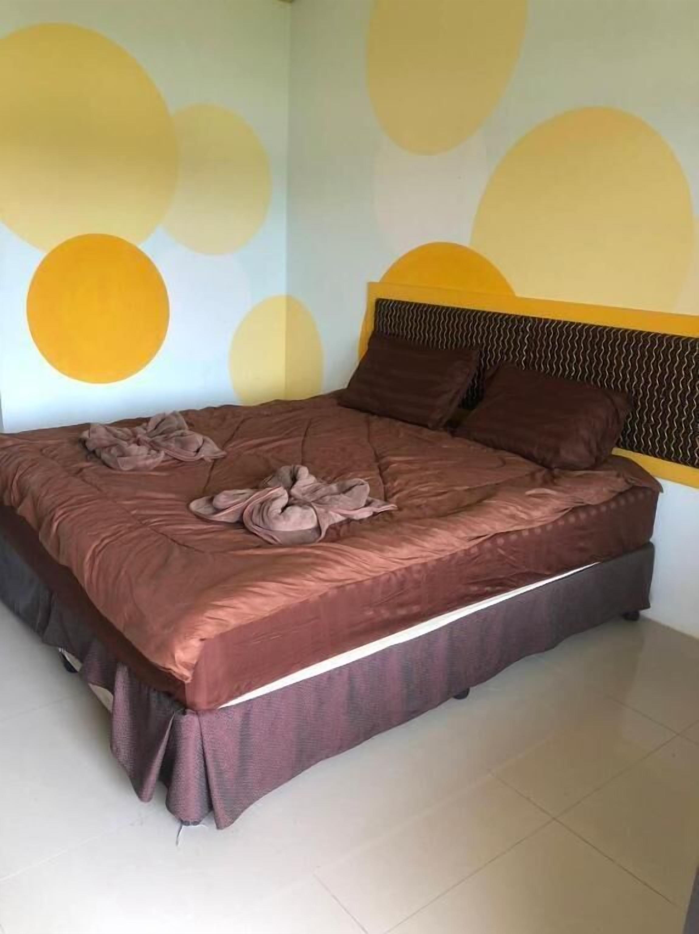 Een bed met een bruine sprei en twee kussens, een hoofdbord met een golvend patroon, en een muur met grote gele cirkels.