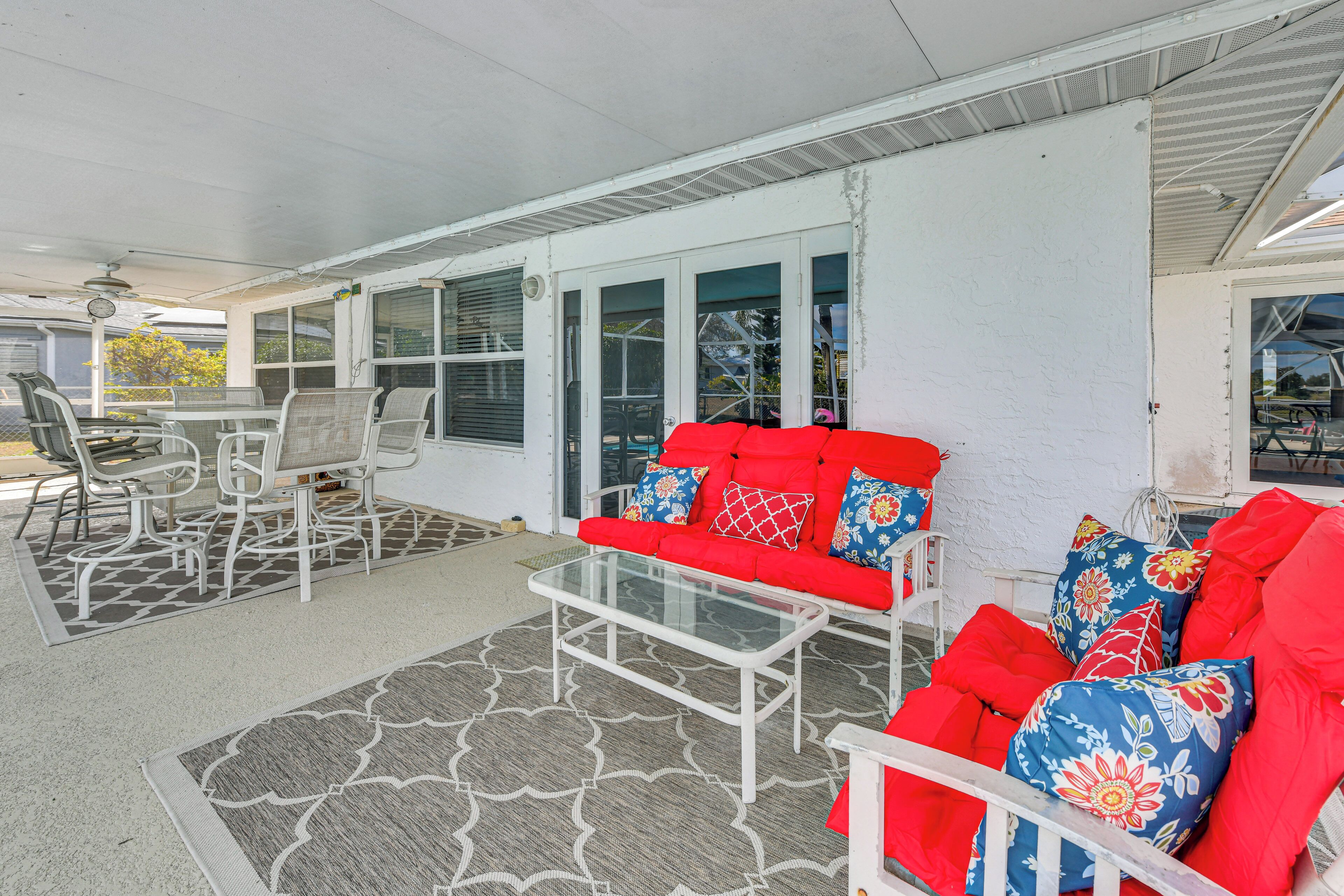 A covered patio area with a white ceiling, a glass coffee table, a red sofa with floral cushions, and a dining table with chairs.