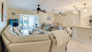 A living room with a beige sofa, a coffee table, and a TV. There are ceiling fans, a kitchen area with cabinets, and a door leading to another room.