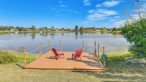 A wooden dock with two red Adirondack chairs facing a calm lake, surrounded by greenery and a clear blue sky.