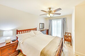A bedroom with a bed, wooden nightstand, dresser, ceiling fan, and a window with curtains. - Patio & Workspace: Peaceful Riverside Retreat! (Riverside)