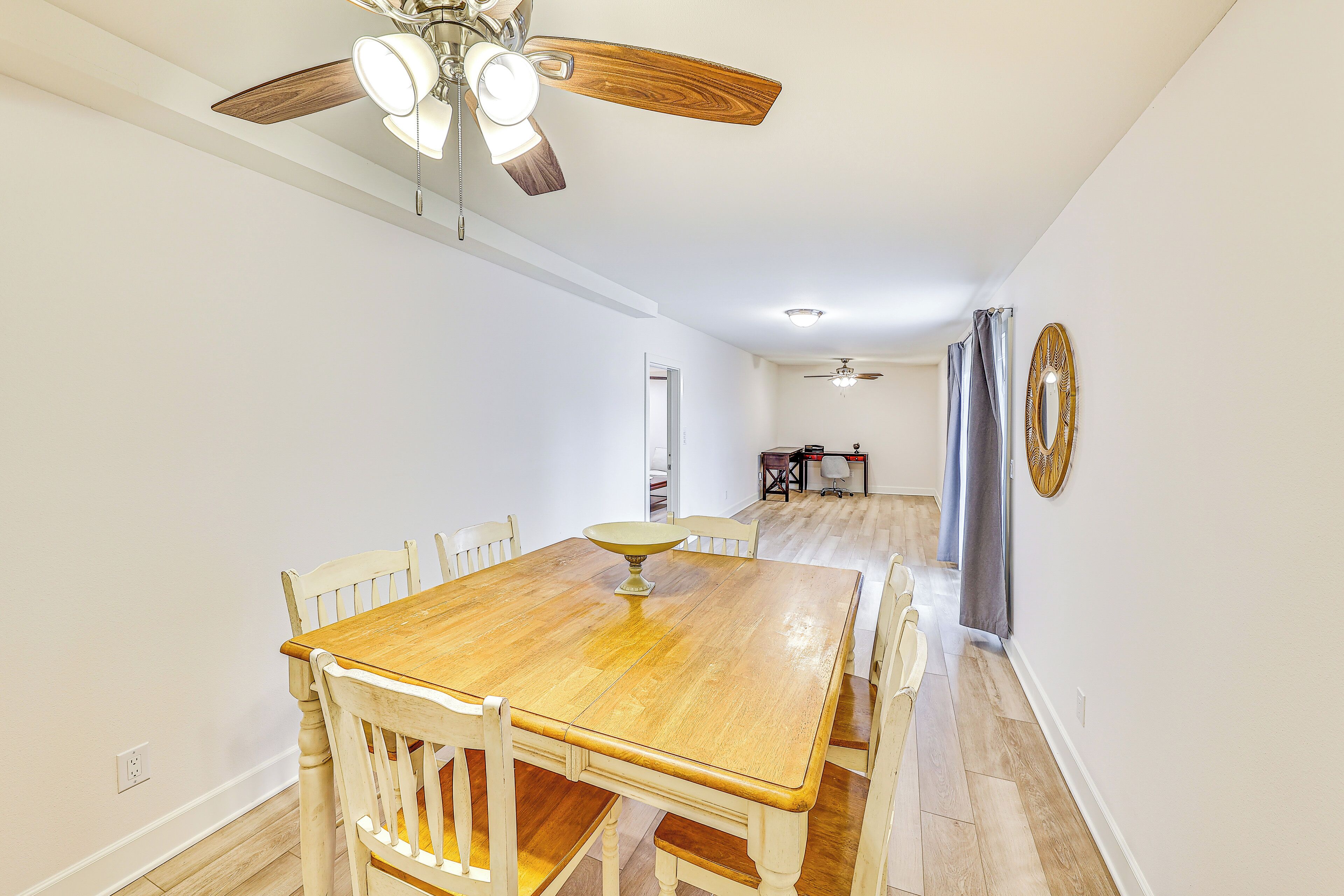 Un coin repas avec une table et des chaises en bois, un ventilateur de plafond et un couloir menant à une autre pièce.