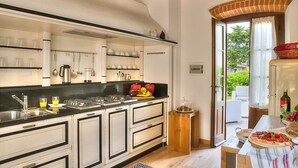 A kitchen with white cabinets, black countertops, and a wooden ceiling. There is a dining area with a table set for two, a window with curtains, and a view of greenery outside.