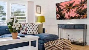 A living room with a blue sofa, a white coffee table, a patterned cushion, a striped ottoman, a painting of palm trees, and a small table with a lamp. - 4 Br Oasis~Pool~Tiki Hut~Hot Tub~Ping Pong~Arcade (Seminole)