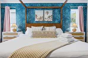 Un lit avec une tête de lit, deux tables de chevet, un mur orné d’un motif de feuilles tropicales et deux tableaux encadrés.